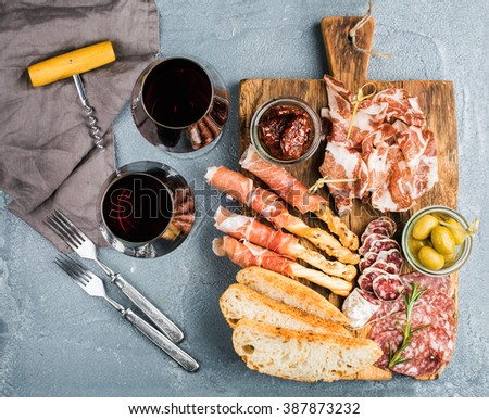 Meat appetizer selection. Prosciutto di Parma, salami, bread sticks, baguette slices, olives, sun-dried tomatoes on rustic wooden board, two glasses of red wine over grey concrete textured backdrop