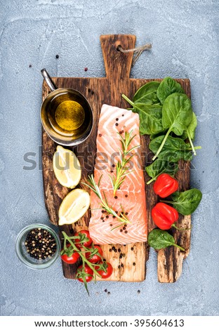 Ingredients for cooking healthy dinner. Raw salmon fillet, spinach, tomatoes, olive oil, lemon, peppers, rosemary and spices on a rustic wooden board over concrete textured grey background. Top view