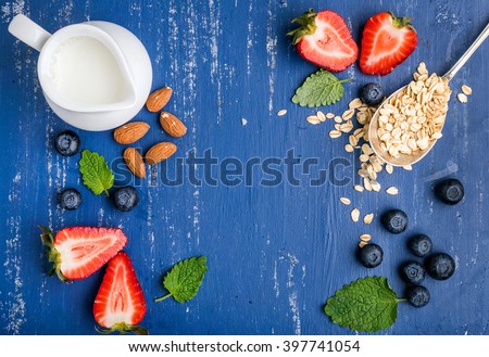 Healthy breakfast ingredients food frame. Oatmeal in spoon, milk in creamer, berries, almond and mint on painted blue wooden background, top view, copy space