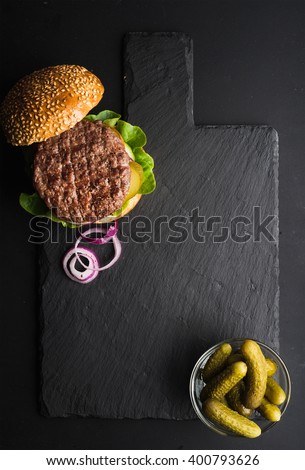 Fresh homemade burger on dark slate stone board, pickles and sliced onion over black background. Top view, copy space
