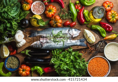 Raw uncooked seabass fish with vegetables, grains, herbs and spices on chopping board over rustic wooden background, top view