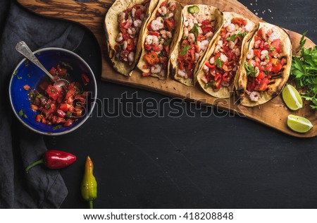 Shrimp tacos with homemade salsa, limes and parsley on wooden board over dark background. Top view, copy space. Mexican cuisine