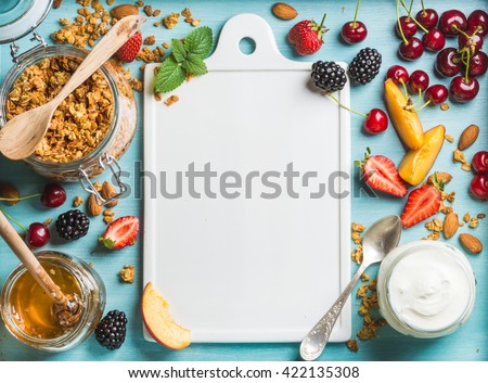 Healthy breakfast ingredients. Oat granola in open glass jar, yogurt, fruit, berries, honey and mint on blue background with white ceramic board in center, top view, copy space