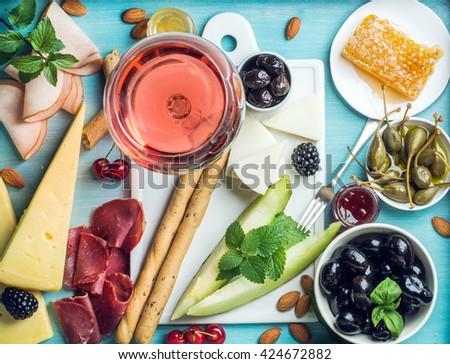Summer wine snack set. Glass of rose, meat, cheese, olives, honey, bread sticks, nuts, capers and berries with white ceramic board in center, blue wooden background, top view