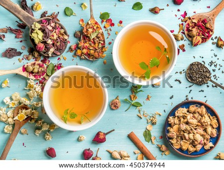 Two cups of healthy herbal tea with mint, cinnamon, dried rose and camomile flowers in spoons over blue background, top view
