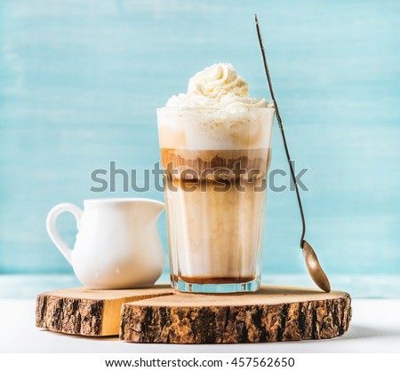 Latte macchiato with whipped cream, serving silver spoon and pitcher on wooden round board over blue painted wall background, selective focus, horizontal composition