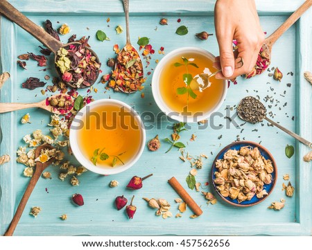 Two cups of healthy herbal tea with mint, cinnamon, dried rose and chamomile flowers in spoons and man\'s hand holding spoon of honey, blue background, top view