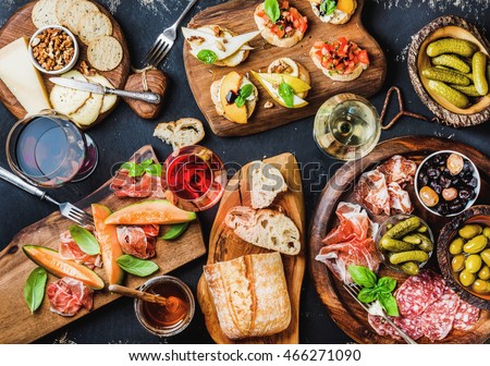 Italian antipasti wine snacks set. Brushettas, cheese variety, Mediterranean olives, pickles, Prosciutto di Parma with melon, salami and wine in glasses over black grunge background, top view