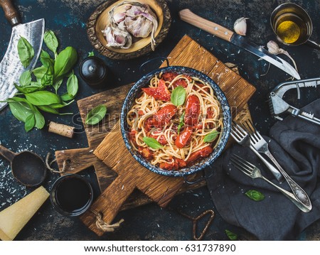 Italian style pasta dinner. Spaghetti with tomato and basil in plate on wooden board and ingredients for cooking pasta over dark plywood background, top view