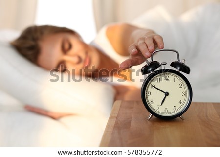 Sleepy young woman stretching hand to ringing alarm to turn it off. Early wake up, not getting enough sleep, getting work concept.
