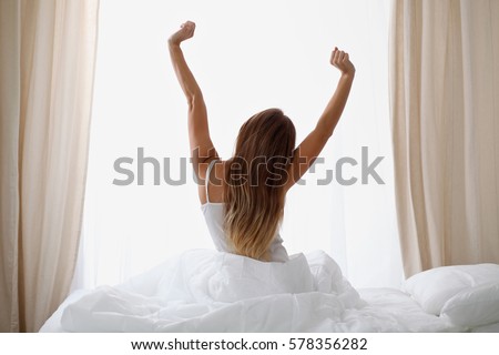Woman stretching in bed after waking up, back view, entering a day happy and relaxed after good night sleep. Sweet dreams, good morning, new day, weekend, holidays concept