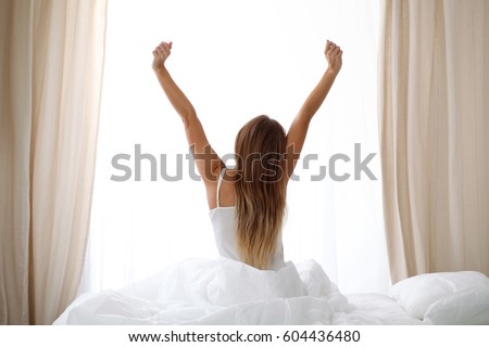 Alarm clock standing on bedside table has already rung early morning to wake up woman is stretching in bed in background. Early awakening, not getting enough sleep, oversleep concept
