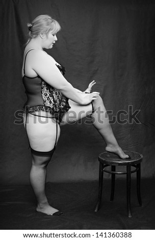 Vintage style picture of an overweight woman dressed in black lingerie posing on a black background.