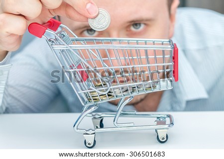 Man throws a coin into money box of the shape of trolley. The conception of how to cost-effective spend money while shopping.