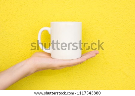 Young Caucasian Woman Holds on Hand Palm Blank Mockup White Mug on Bright Yellow Painted Wall. Airy Breezy Style. Template for Text Artwork Lettering. Trendy Minimalist Urban Atmosphere