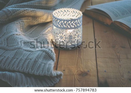 Lit Candle White Knitted Sweater Open Book on Plank Wood Table by Window. Cozy Winter Autumn Evening. Natural Light Authentic Tranquil Atmosphere. Kinfolk Hygge Slow Living Style. Matte Toned