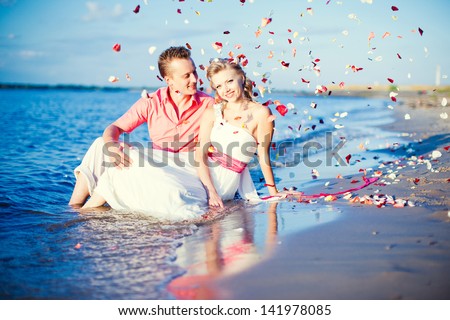 The bride and groom in their wedding clothes lie in the water of the sea. Trash the dress. After the wedding. Fun, romance. Newlyweds lie on the beach. falling rose petals.