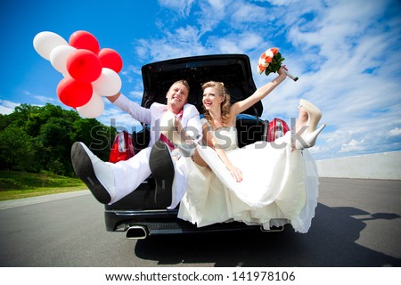 Happy Beautiful Bride And Groom Sitting In The Trunk Of A Car. Honeymoon, Fun And Laughter.