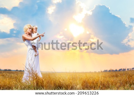 Happy family in a meadow. Mother holds child on hands on a background of a beautiful sunset in the field. Woman points a finger in the free space on the photo.