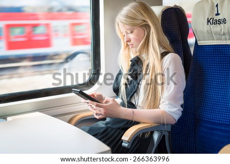 Blond girl sitting in First Class of a train and holding a tablet in her hands. On the sit is written \