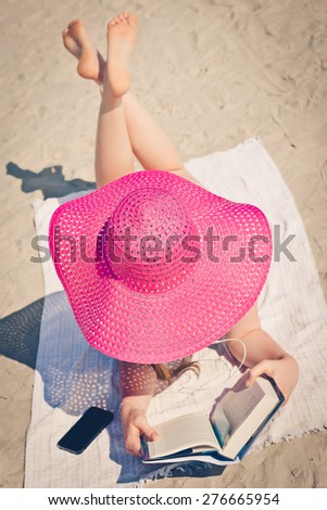 Girl in a big pink hat relaxing on beach, reading a book and listening to music from her smart phone. Selective focus. Top view.