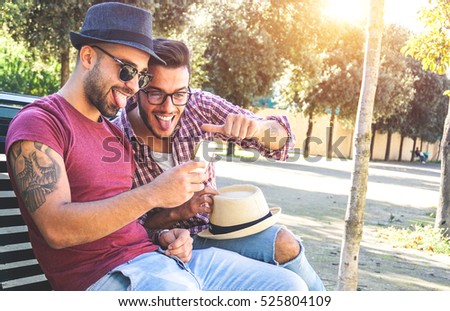 Two handsome friends taking a selfie with a smart phone sitting on a bench on a sunny day looking the camera making funny face