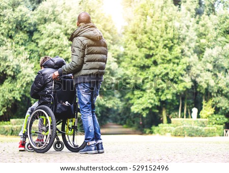 Back view of a carer man with young disabled with handicap on wheelchair outdoor in a park - Special needs care assistant working with disability - Social issues with invalid guy on difficulties