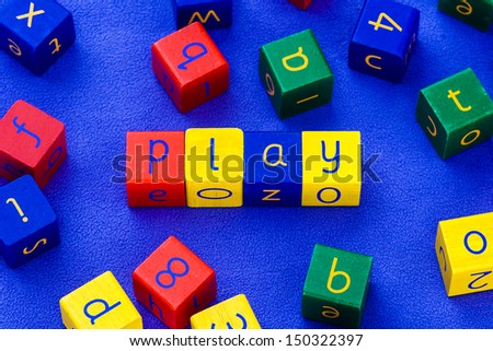 The word 'PLAY', written with colored, wooden toy blocks. Arranged on a blue, soft carpet.