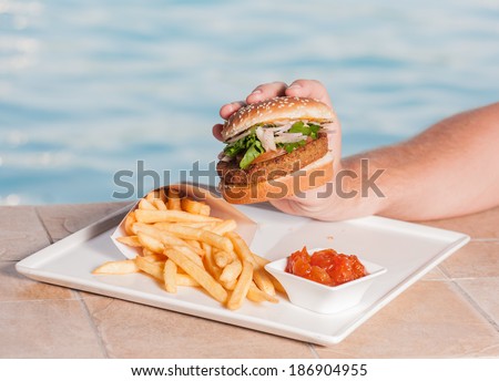Hand holding burger with fries and sauce,swimming pool in background