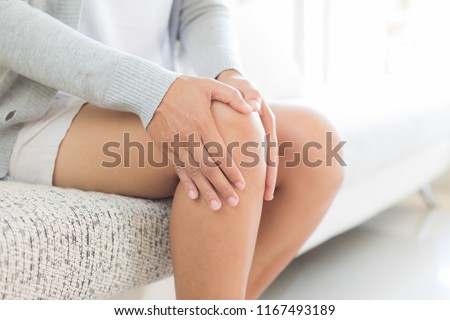 Closeup young woman sitting on sofa and feeling knee pain and she massage her knee at home. Healthcare and medical concept.