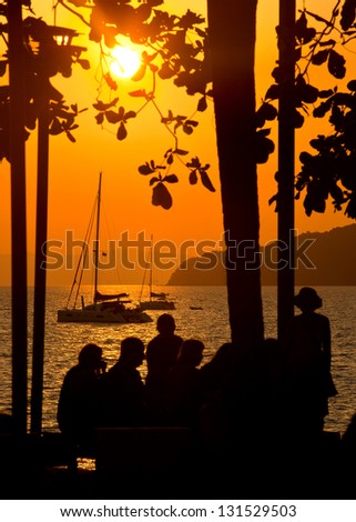 A group of people on the beach looking at the sailing yacht, illuminated by the light of the sun setting
