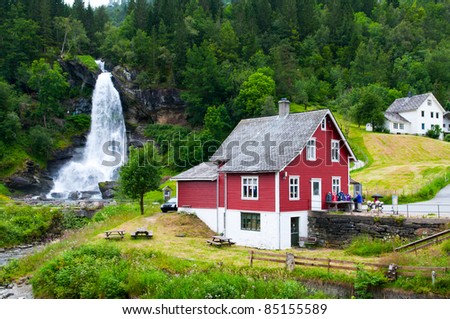 Traditional norwegian red wooden house with waterfall in the distance. Waterfall Steindalsfossen, Norway.