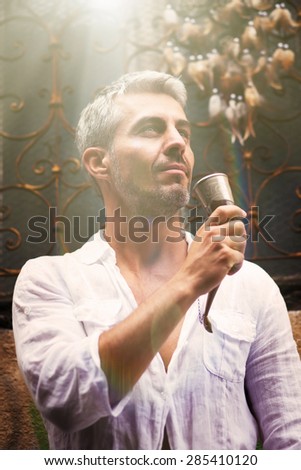 Sexi Man in a white shirt and medieval mead horn in hand. Toast concept. Ornamental window on background, and Dream Catcher. in sun light