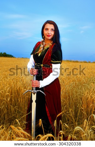 Smiling Young woman with ornamental dress and sword in hand  standing on a wheat field with sunset. Natural background.