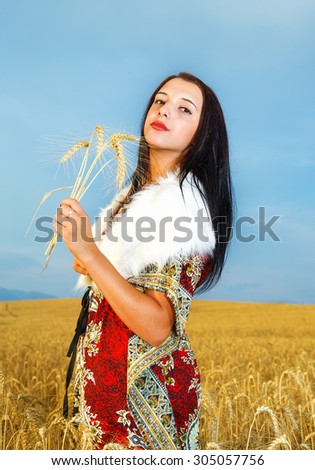 Young woman with ornamental dress and white fur standing on a wheat field with sunset. Holding bouquet
