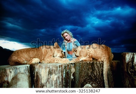 sexy blonde woman playing with lion cub on background with beautiful blue sky and storm clouds