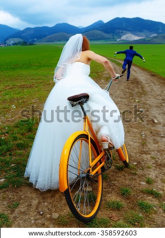 bride on orange retro bike is chasing after a groom in blue wedding suit with a beer bottle. wedding concept.