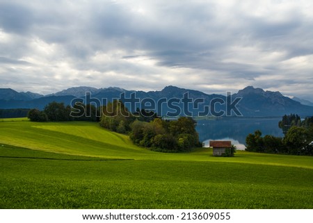 Beautiful landscape - green hill with trees and house on the lake in Bavaria, with the Alps mountains in the background, Germany, cloudy summer day