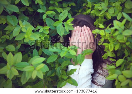 Girl hiding in the green bushes, covering her face with both hands. Outdoors