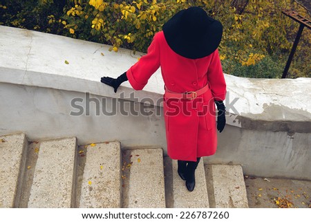 Young woman hiding her face under black hat, standing on the stairs. Autumn bushes background. Top view