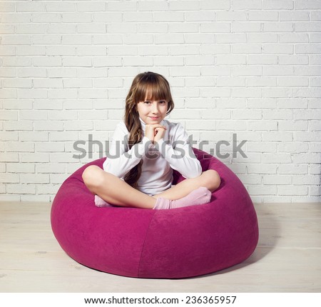 Young pretty girl sitting on pink padded stool against white brick wall. Studio photo