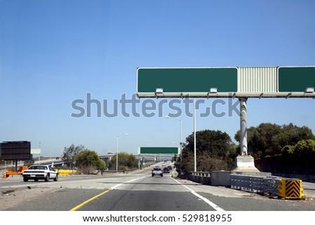 Passenger view of two blank interstate exit signs under blue sky. One car. Little traffic. Trees on horizon with blue sky. Horizontal.