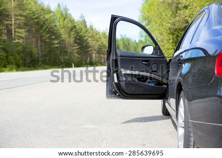 A black vehicle with an open door in side of the road. Image taken during summer. The driver is needing for help or giving assistance for example.