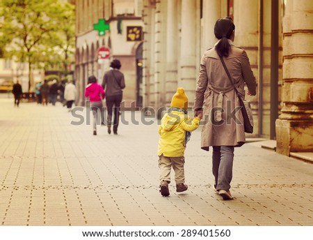 A brown haired is mom taking a child to a kindergarten. The boy is jumping happily on the way.