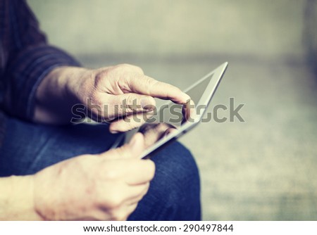 An elderly man browsing the internet with a tablet in a living room and sitting in a sofa. Image has a vintage effect.
