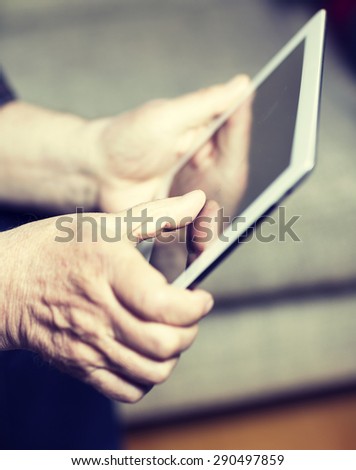 An elderly man browsing the internet with a tablet in a living room and sitting in a sofa. Image has a vintage effect.
