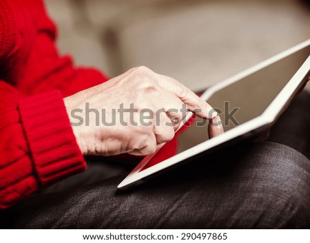 An elderly woman browsing the internet with a tablet in a living room and sitting in a sofa. Image has a vintage effect.