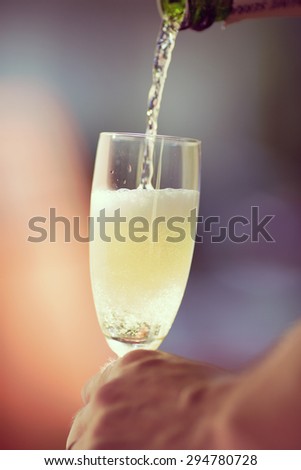 An image of someone pouring champagne to a glass in the summer and outdoors. Image has a vintage effect.
