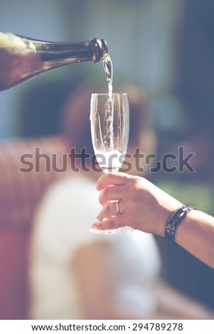 An image of pouring champagne to a glass outdoor and in the summer time. Image has a vintage effect applied.