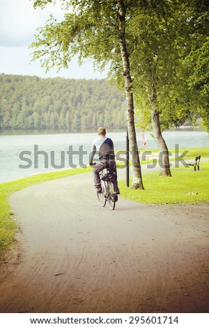 A man riding a bike in the park next to a lake. Some forest is on the other side of the lake and the curve is turning right. Image has a vintage effect applied.